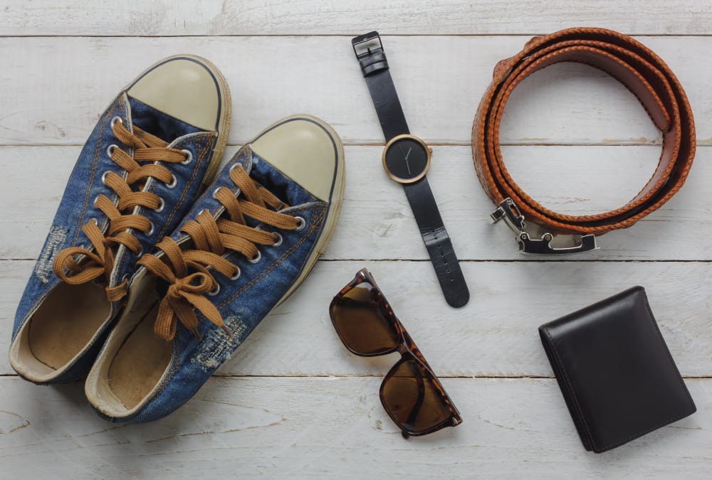 Acessórios masculinos organizados sobre uma superfície de madeira, incluindo tênis casuais, relógio de pulso, cinto de couro, óculos de sol e carteira.